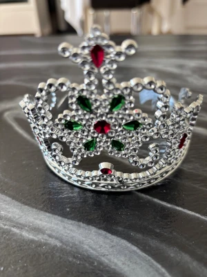 Glittrande tiara med röda och gröna stenar - Säljer en snygg tiara i silverfärgad metall dekorerad med glittrande röda och gröna plaststenar. Kronan har en klassisk form med detaljerade mönster och gnistrande yta som verkligen sticker ut. Perfekt för dig som vill känna dig extra kunglig.