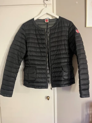 Svart pufferjacka från Colmar - Snygg svart pufferjacka från Colmar med rund hals, två framfickor och tydlig logga på ärmen. Jackan har en silverfärgad dragkedja med Colmar-detalj. Perfekt för dig som gillar stilren och sportig look.