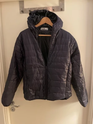 Mörkblå pufferjacka från Stone Island - Säljer en mörkblå pufferjacka från Stone Island med huva och dragkedja framtill. Jackan har design och klassisk Stone Island patch på vänster ärm. Perfekt för kyliga dagar och har en clean, sportig look. Jackan är i storlek S. Fick den i present men säljer den nu på grund av att den är för liten. Jackan är mer eller mindre i nyskick då den endast är testad ett fåtal gånger.