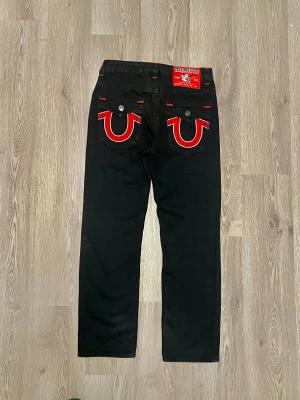 True Religion svarta jeans med röda detaljer  - Svarta jeans från True Religion, modell Joey Super T. Använde dom 2-3 gånger. Snygga kontrastsömmar i rött och silverfärgade knappar. Bakfickorna har ikonisk broderad logga i rött och vitt. Klassisk rak passform och robust jeansmaterial. Perfekt för dig som gillar streetwear med attityd.   Längd: 112 cm Midja: 42 Raise: 27 Lår: 26