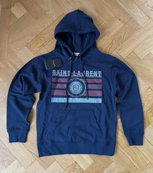Marinblå hoodie Saint Laurent Paris - Snygg marinblå hoodie från Saint Laurent Paris med tryck i rött, vitt och blått framtill. Klassisk känguruficka, dragsko i huvan och ribbade muddar. Mjuk bomullskvalitet och cool vintageinspirerad print med SL-emblem. Perfekt för en chill och trendig look.