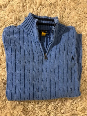 Blå stickad polotröja från Polo Ralph Lauren - Snygg blå polotröja från Polo Ralph Lauren med klassiskt kabelstickat mönster och halv dragkedja vid halsen. Tröjan har ribbade muddar och lång ärm, samt den ikoniska Polo-loggan broderad på bröstet. Perfekt för dig som gillar stilren och tidlös design. Det är storlek XL barn 