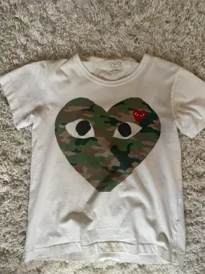 Säljer en vit t-shirt från Comme des Garçons Play i storlek S. Framsidan har ett stort hjärta med camouflage-mönster och ett mindre rött broderat hjärta med ögon. T-Shirten sitter mindre i storleken så mer S-XS. Hör av dig vid frågor eller fler bilder! 