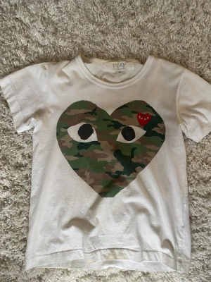 Vit Comme des Garçons Play t-shirt S - Säljer en vit t-shirt från Comme des Garçons Play i storlek S. Framsidan har ett stort hjärta med camouflage-mönster och ett mindre rött broderat hjärta med ögon. T-Shirten sitter mindre i storleken så mer S-XS. Hör av dig vid frågor eller fler bilder! 