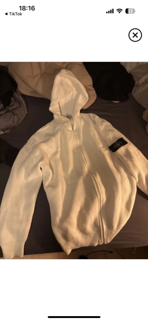 Vit stickad hoodie från Stone Island - Säljer en vit stickad hoodie från Stone Island med dragkedja framtill och klassisk patch på vänster ärm. Jackan har ribbad struktur och huva, perfekt för dig som gillar clean och stilren streetwear.