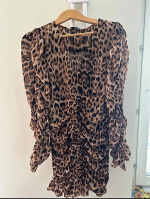 Leopardmönstrad kortklänning NA-KD - Säljer en superfin kort klänning från NA-KD i leopardmönster. Klänningen har v-ringning, långa puffiga ärmar och snygga rynkade detaljer framtill. Materialet är tunt och lätt, perfekt för att sticka ut. Färgerna går i beige, brun och svart.