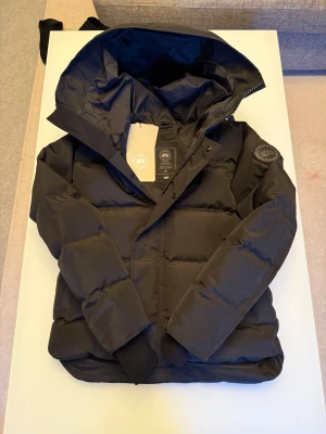 Svart pufferjacka från Canada Goose - Svart pufferjacka från Canada Goose i storlek M. Jackan har en stor huva, quiltad design och logotyp-patch på ärmen. Materialet är polyester och dun, perfekt för kalla dagar. Stängs med dragkedja och tryckknappar framtill. Snygg och funktionell för vintern.