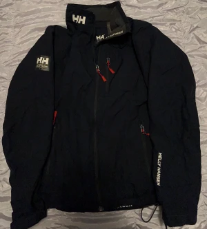 Svart vindjacka Helly Hansen M - Svart vindjacka från Helly Hansen med Helly Tech-skydd, röda detaljer på dragkedjorna och logga på ärm och krage. Jackan har hög krage, meshfoder och flera fickor med dragkedja. Perfekt för blåsiga dagar och outdoor.