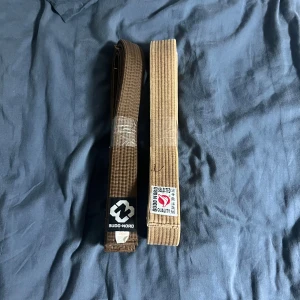 Brunt Budo-Nord bälte 300cm - Två bruna kampsportsbälten från Budo-Nord, båda 300 cm långa. Bältena är tillverkade i bomull och polyester och har klassiska broderade etiketter med japanska tecken och logga. Perfekt för dig som tränar judo eller karate och vill ha ett stilrent, klassiskt bälte.