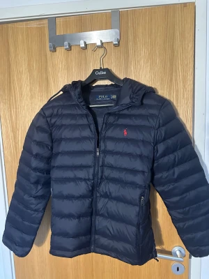 Ralph Lauren jacka - Snygg mörkblå dunjacka från Polo Ralph Lauren med huva och dragkedja. Jackan har quiltad design, två fickor med dragkedja och klassisk röd logga på bröstet. Perfekt för kyliga dagar och enkel att matcha med olika outfits.