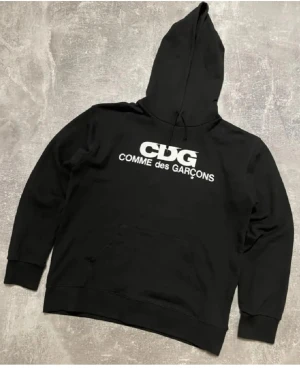 Svart hoodie från Comme des Garçons - Svart hoodie från Comme des Garçons med stor vit CDG-logga på bröstet. Tröjan har huva med snörning, känguruficka och långa ärmar. Perfekt för dig som gillar streetwear och vill ha en clean look med ikonisk branding.