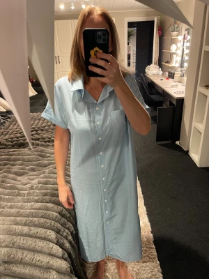 Ljusblå skjortklänning med slits - Säljer en ljusblå skjortklänning med knappar hela vägen framtill, klassisk krage och korta ärmar med uppvik. Klänningen har en slits på sidan och en bröstficka. Perfekt för dig som gillar enkel och chill stil.