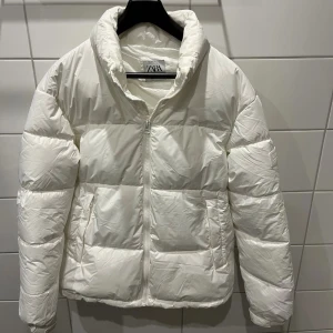 Vit pufferjacka från Zara, storlek M - Snygg vit pufferjacka från Zara i storlek M. Jackan har hög krage, dragkedja framtill och två fickor på sidorna. Perfekt för kyliga dagar och ger en clean look. Tillverkad i syntetmaterial och har en klassisk pufferdesign.