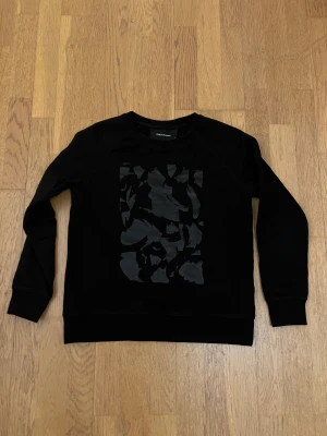 Svart sweatshirt från Peak Performance - Svart sweatshirt från Peak Performance i storlek S. Tröjan har ett coolt ton-i-ton tryck på bröstet och är långärmad med ribbade muddar vid ärmslut och nederkant. Perfekt för dig som gillar stilrena och sportiga plagg.