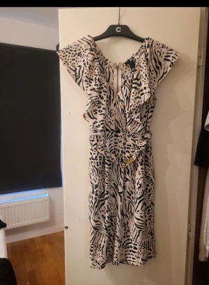 Zebramönstrad kortklänning från H&M - Säljer en kortklänning från H&M med zebramönster i svart och beige. Klänningen har volangdetaljer vid axlarna, v-ringad hals och ett brett midjebälte med guldspänne. Materialet är lätt polyester och klänningen har dragkedja i ryggen.