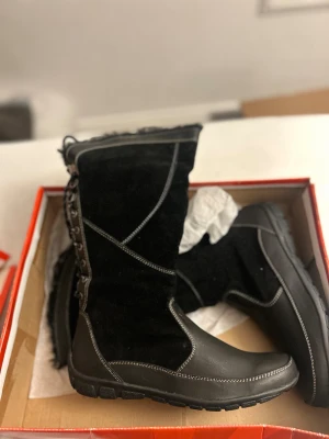 Svarta vinterstövlar med snörning från Skills, finns i storlek 39 och 40 - Svarta stövlar i mocka och skinn med grov sula och dekorativa kontrastsömmar. Stövlarna har snörning baktill, dragkedja på sidan och mjukt foder upptill. Perfekta för kalla dagar och har en klassisk, stilren look. Päls i insidan, finns i storlek 39 och 40.