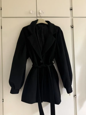 Svart kappa med bälte och knappar - Stilren svart kappa med breda slag, knappar framtill och ett matchande bälte i midjan. Jackan har långärmade puffärmar med knappdetalj vid ärmslutet. Perfekt för dig som gillar en klassisk look med modern twist.