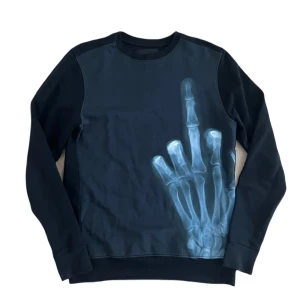 Svart sweatshirt Limitato - Cool svart sweatshirt från Limitato med ett unikt röntgenmotiv av en hand som visar långfingret på framsidan. Tröjan har rund hals, långa ärmar och är tillverkad i mjuk bomull. Perfekt statement piece för dig som vill sticka ut.