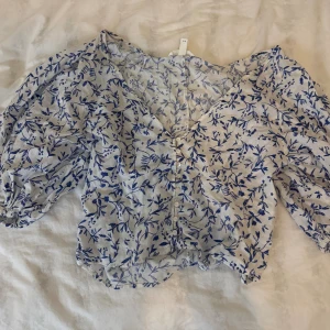 Blommig blus med puffärm - Säljer en söt blommig blus från H&Mi vit och blå. Blusen har puffiga trekvartsärmar, v-ringning och små knappar framtill. Materialet är lätt och luftigt, perfekt för varma dagar. Blommönstret ger en fräsch och somrig vibe.