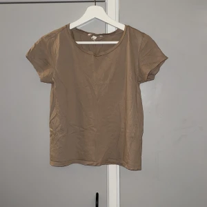 Beige kortärmad topp från H&M - En enkel och stilren beige topp från H&M i storlek XS. Modellen är kortärmad med rund halsringning och rak passform. Tillverkad i mjuk bomull som känns skön mot huden. Perfekt att matcha med jeans eller kjol för en clean look.