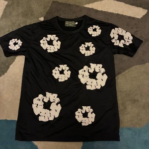 Svart t-shirt med vita tryck från Denim Tears - Svart t-shirt från Denim Tears med unika vita grafiska tryck i cirkelform över hela plagget. T-shirten har rund hals och korta ärmar, tillverkad i mjuk bomull som känns skön mot huden. Perfekt för dig som gillar streetwear och vill sticka ut.