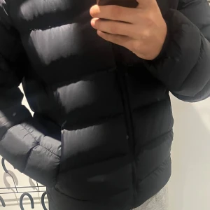 Svart pufferjacka från peak performance - Säljer en svart pufferjacka från peak performance med diskret logga på ärmen. Jackan har en stilren, quiltad design och dragkedja framtill. Den är långärmad och har en normal passform, perfekt för kalla dagar. Fylld med syntetmaterial för extra värme.