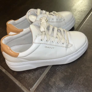 Vita sneakers från GANT med beige detalj - Snygga vita sneakers från GANT med beige hälparti och klassisk rund tå. Skorna har snörning och är tillverkade i skinn med platt sula. Perfekta för dig som gillar stilrena och fräscha sneakers med en liten twist.