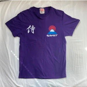 Samurai Jeans t-shirt - Fet lila t-shirt från Samurai Club med japanska tecken och coolt tryck av Mount Fuji och sol på både fram- och baksidan. T-shirten har rund hals och korta ärmar, tillverkad i mjuk bomull. 