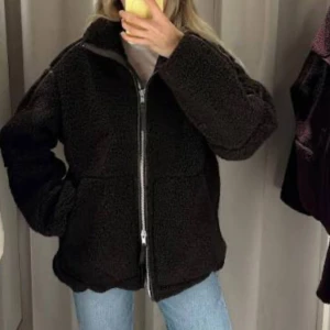Svart teddyjacka med dragkedja - Mysig svart teddyjacka med hög krage och hel dragkedja framtill. Jackan har en loose passform och är tillverkad i mjukt fleece-material som håller dig varm. Perfekt att slänga på sig när det är kyligt ute. Snygg och enkel att matcha med jeans eller leggings.