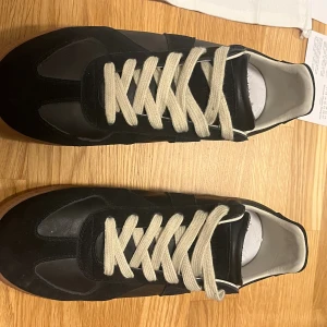Maison Margiela svarta sneakers - Maison Margiela sneakers i svart med kontrasterande vita snören och vit insida. Klassisk rund tå och platt sula. Materialmix av skinn och mocka ger en lyxig känsla. Levereras med originalkartong, dustbag (allt og). Perfekt för dig som gillar stilrena och exklusiva sneakers. Storlek 45