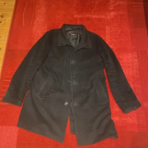 Svart rock från Oscar Jacobson, strl 54 - Svart rock från Oscar Jacobson, modell Storviker Coat. Klassisk design med krage, knäppning framtill och två snedställda fickor. Yttermaterialet har en mjuk känsla och insidan är fodrad med slätt tyg. Perfekt för dig som gillar stilrena och tidlösa plagg. Finns ett hål på höger sidan av fickan dock.