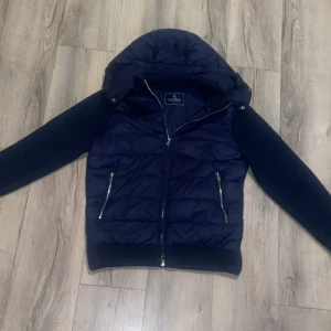 Mörkblå pufferjacka från Vonroe - Snygg mörkblå pufferjacka från Vonroe med stickade ärmar och huva. Jackan har två silvriga dragkedjefickor fram och quiltad front. Perfekt för dig som vill ha en cool och bekväm look under höst och vinter.