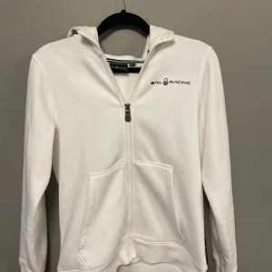Vit hoodie från Sail Racing med dragkedja - Vit hoodie från Sail Racing med svart logga på bröstet och huvan. Tröjan har hel dragkedja framtill, två stora fickor och lång ärm. Mjuk bomullskänsla och klassisk passform, perfekt för chill dagar. Snygg och enkel design med coolt broderi på huvan.