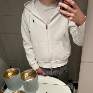 Vit kofta från Polo Ralph Lauren - Vit kofta från Polo Ralph Lauren med dragkedja och snörning i huvan. Klassisk svart logga på bröstet, två fickor framtill och ribbade muddar. Den har endast använts 2-3 gånger så den är så gott som ny. Jättesnygg kofta som passar till nästan allt. OBS Jag la ut denna för nån månad sen men imed att den returnerades tillbaka till mig för köparen ej kunde genomföra uthämtning osv. Hör av er om ni har frågor!
