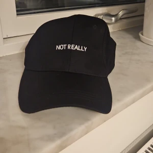 Svart keps med texten 'Not Really' - Svart keps med böjd skärm och vit broderad text 'NOT REALLY' framtill. Justerbar rem med metallspänne baktill för perfekt passform. Tillverkad i mjukt bomullstyg, enkel och stilren design som passar till streetwear och chill vardagsstil.