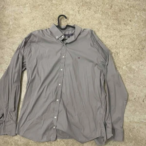 Grå skjorta från Tommy Hilfiger - Snygg grå skjorta från Tommy Hilfiger med klassisk krage och diskret logga på bröstet. Skjortan har långa ärmar, knappar framtill och är tillverkad i mjuk bomull. Perfekt för dig som gillar stilrena och enkla plagg.