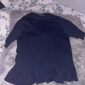 Mörkblå topp med volang och ribbat  - Snygg mörkblå topp  med smockad front och volangkant nedtill. Tröjan  har rund hals och korta ärmar, tillverkad i mjukt bomullstyg. Perfekt för dig som gillar enkel stil med en twist.