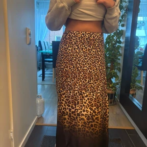 Leopardmönstrad långkjol i satin - Säljer en snygg långkjol med leopardmönster och mörkare toning nedtill. Kjolen har en rak och flowy passform som sitter skönt i midjan. Materialet är glansigt satin som ger en lyxig känsla och cool look. Perfekt för dig som vill sticka ut med djurmönster.