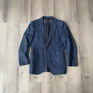 Blå kavaj från Suitsupply - Snygg blå kavaj från Suitsupply, modell Lazio, i 100% ullflanell från Vitale barberis canonico med ett diskret rutigt mönster. Kavajen har två knappar framtill, fyra knappar vid det öppningsbara ärmslutet och en klassisk passform. 