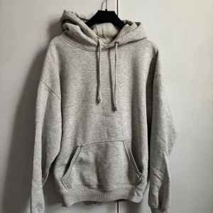 Grå hoodie med huva och magficka - En klassisk grå hoodie med huva och snörning, samt en stor magficka framtill. Tröjan har ribbade muddar vid ärmslut och nederkant, och är tillverkad i mjukt material som passar perfekt för chill dagar.