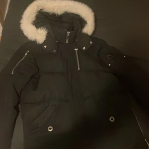 Svart Moose Knuckles dunjacka med päls - Säljer en svart dunjacka från Moose Knuckles med vit fuskpäls runt huvan. Jackan har flera dragkedjor, snygga metalldetaljer och logga på ärmen. Fodrad med dun för extra värme och har en cool broderad insida. Perfekt för kalla vinterdagar.