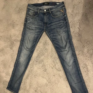 Replay Anbass jeans blå 33/32 - Snygga Replay Anbass jeans i klassisk blå tvätt med diskreta slitningar och kontrastsömmar. Modellen har fem fickor och läderpatch bak i midjan. Perfekt för dig som gillar stilrena och moderna jeans med lite edge.