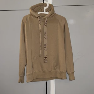 Beige hoodie från Zink med logoband - Säljer en beige hoodie från Zink i storlek M. Tröjan har huva, långa ärmar och en stor magficka. Snygga breda snören med Zink-logga ger en cool detalj. Perfekt för chill dagar eller när du vill ha en enkel men stilren look.