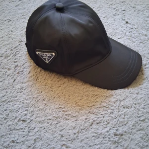 Svart Prada keps i nylon - Snygg svart keps från Prada med klassisk böjd skärm och ikonisk metalltriangel-logga på sidan. Tillverkad i återvunnen nylon som ger en stilren och modern känsla. Perfekt accessoar för dig som gillar exklusiva märken och minimalistisk design.
