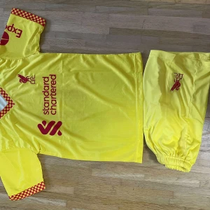 Liverpool Salah gul fotbollströja - Gul Liverpool fotbollströja med M. Salah och nummer 11 på ryggen. Röd logga och tryck framtill, samt röda rutiga detaljer på ärmslut och krage. Tillverkad i lätt polyester, kortärmad och med matchande shorts med LFC-logga.