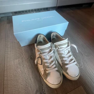 Philippe Model vita sneakers med snörning - Säljer ett par vita Philippe Model sneakers med klassisk snörning och rund tå. Skorna har beige detaljer i mocka och en platt sula i svart och vitt. Märket syns tydligt på plösen och boxen medföljer. Perfekt för dig som gillar stilrena och exklusiva sneakers.