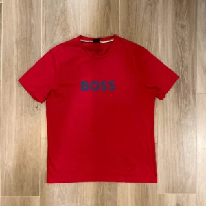 Röd BOSS t-shirt med tryck - Snygg röd t-shirt från BOSS med mörkblått BOSS-tryck på bröstet. Klassisk rund hals och korta ärmar. Perfekt för dig som gillar stilrena och enkla plagg med tydlig logga. Skön bomullskvalitet som känns mjuk mot huden.