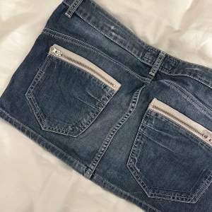 Blå jeanskjol från TRF French - Säljer en blå jeanskjol från TRF French med klassisk femficksdesign, silverfärgade dragkedjor på bakfickorna och knappstängning fram. Kjolen är kort och har en cool, lite tvättad denimlook. Perfekt för dig som gillar streetstyle och denim.