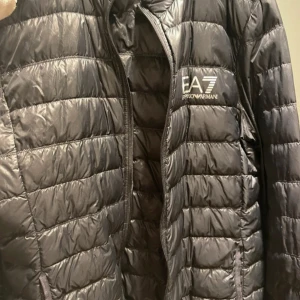Svart pufferjacka från EA7 Armani - Säljer en svart pufferjacka från EA7 Emporio Armani med horisontella quiltade sektioner och dragkedja framtill. Jackan har en stilren look med logotyp på bröstet och är tillverkad i lätt syntetmaterial som passar perfekt för kyliga dagar.