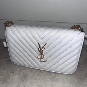 Vit quiltad axelväska från YSL - Supersnygg vit axelväska från YSL med quiltad design och guldiga kedjedetaljer. Väskan har klassisk YSL-logga framtill och är tillverkad i skinn. Perfekt storlek för det viktigaste och ger en lyxig touch till din outfit.